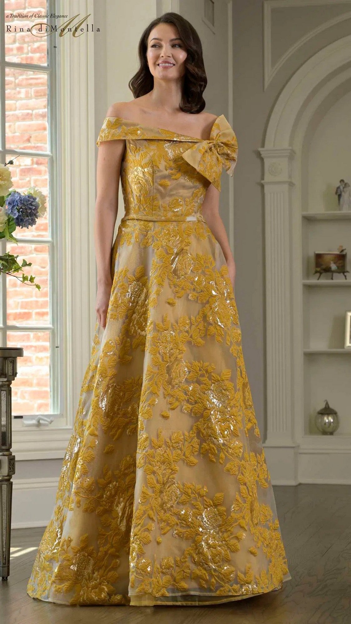 A woman stands indoors wearing the Rina Di Montella RD3401, a gold off-shoulder formal dress with a decorative shoulder bow and an elegant floor-length A-line skirt.